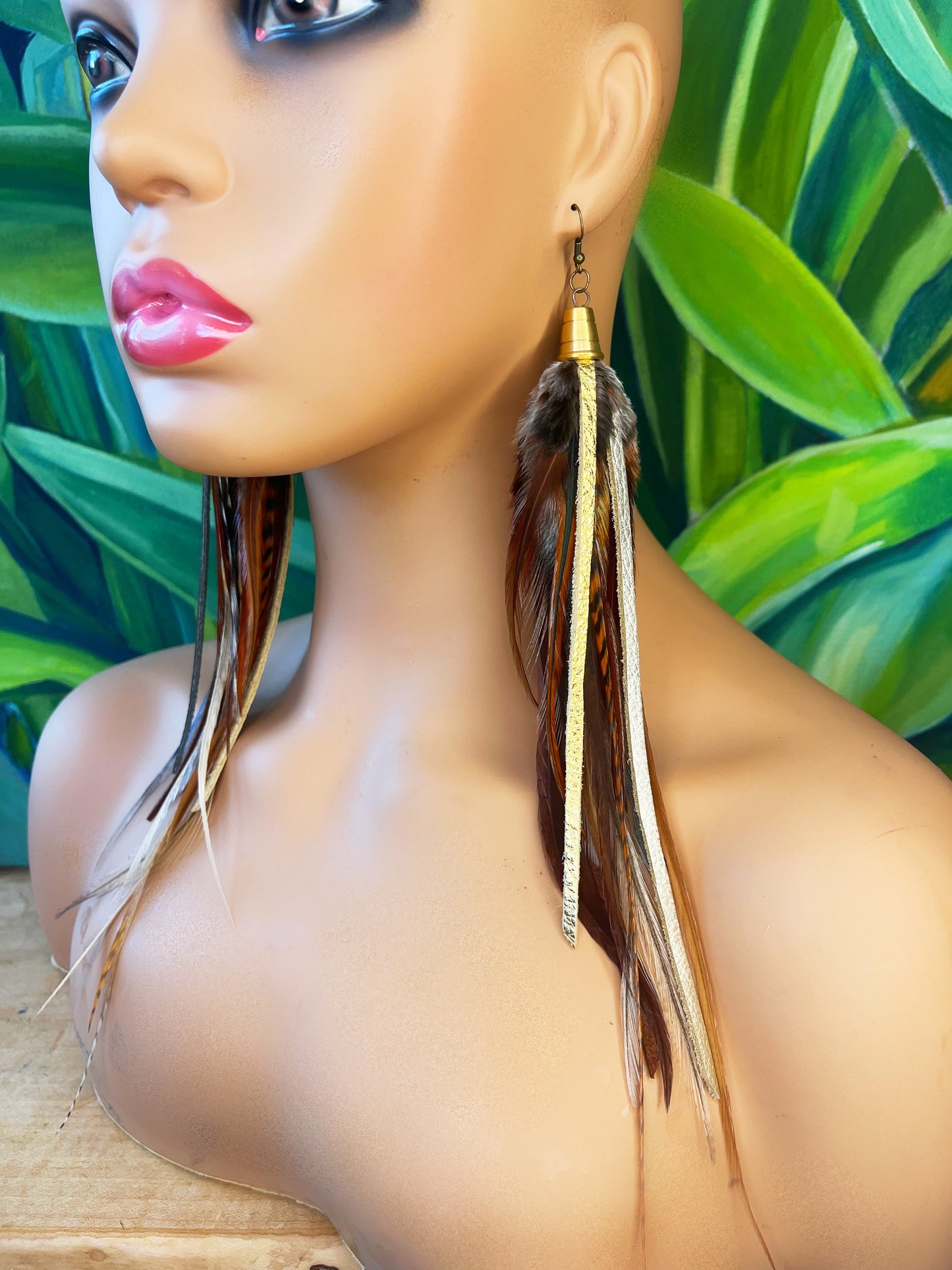 A mannequin head displays a pair of long bohemian earrings, featuring a combination of natural brown feathers and shimmering metallic leather strips in gold, bronze, and pewter. The earrings dangle from gold-colored bronze bead caps connected to hypoallergenic ear wires, set against a lush background of tropical green leaves, which accentuates the natural yet luxurious aesthetic of the jewelry.