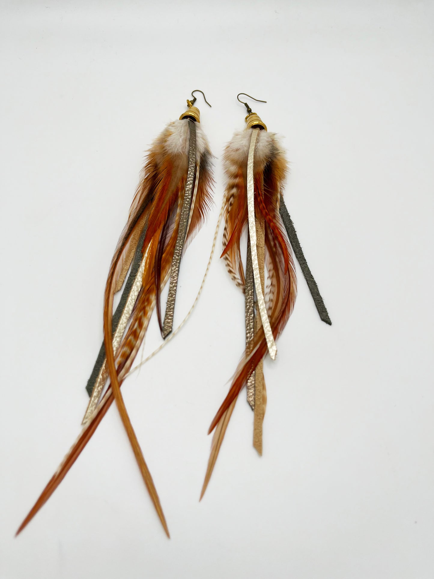 Long feather earrings in light brown and beige, with metallic bronze and pewter colored strips of leather, hanging from a gold colored cap pendant.
