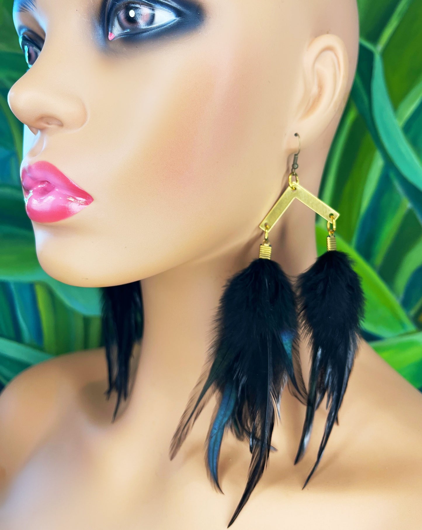A close-up of a mannequin head wearing long black feather earrings. The earrings feature a gold-toned, geometric-shaped hook with a subtle hammered texture, leading to a cascade of sleek black feathers.
