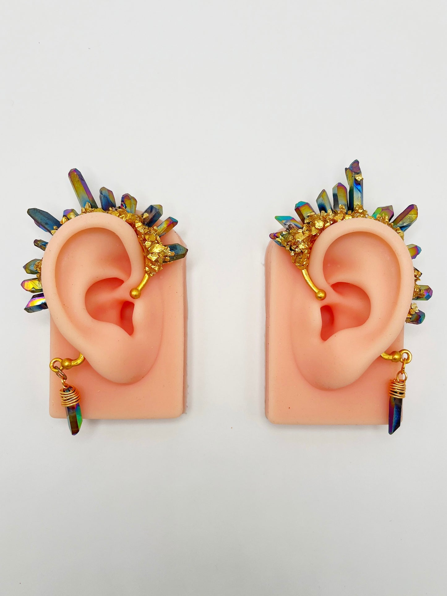A pair of blue rainbow titanium quartz crystal ear cuffs with gold accents, modeled on a pair of ears. A wire wrapped stone hangs from the bottom of each cuff behind the ear.