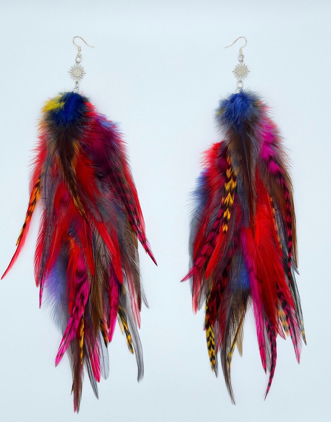 A pair of vibrant feather earrings displayed against a white background. The earrings are adorned with a variety of feathers in bold colors, including red, yellow, blue, and purple, with some natural brown feathers as well. Each earring has a silver fish hook style fastener with a small sunburst patterned charm attached to the top. The feathers are arranged in a cascading, slightly curved fashion, giving the earrings a dynamic and flowing appearance.