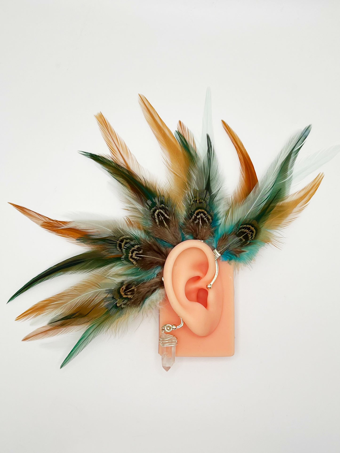 Rooster and pheasant feather ear cuff in green, turquoise and beige tones, modeled on an ear. A wire wrapped quartz stone hangs from the bottom of the cuff behind the ear.