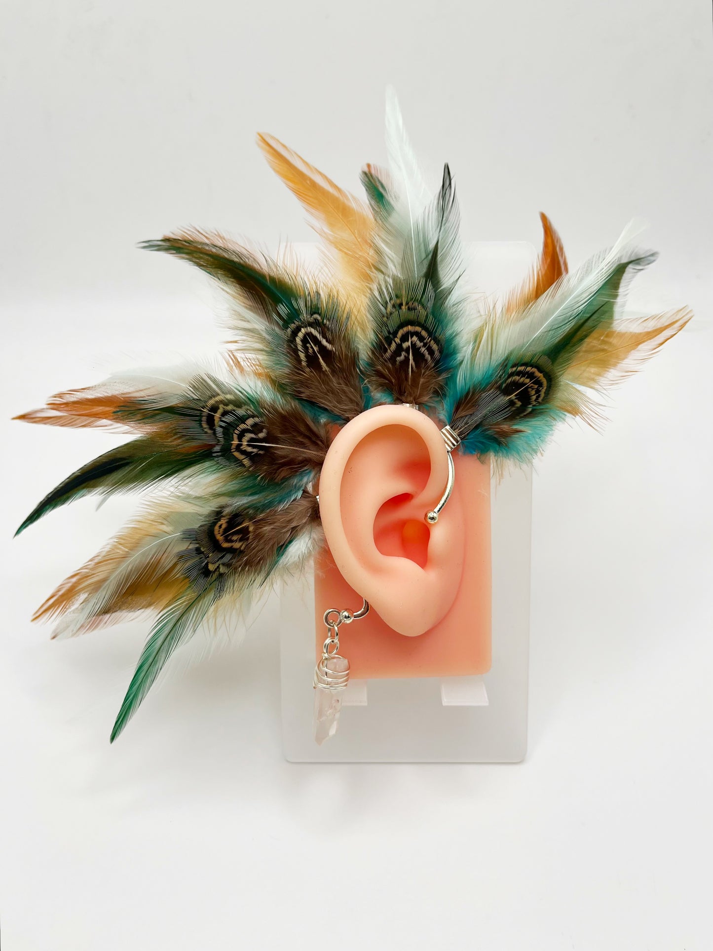 Rooster and pheasant feather ear cuff in green, turquoise and beige tones, modeled on an ear. A wire wrapped quartz stone hangs from the bottom of the cuff behind the ear.