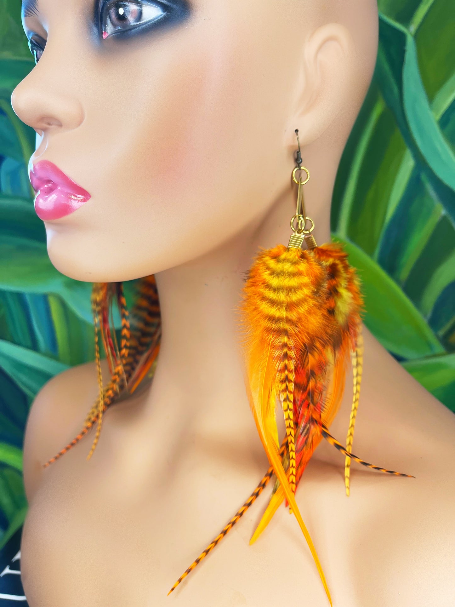 A mannequin wearing medium length bohemian feather earrings in bright golden orange shades, all feathers hang from a gold arrow pendant.