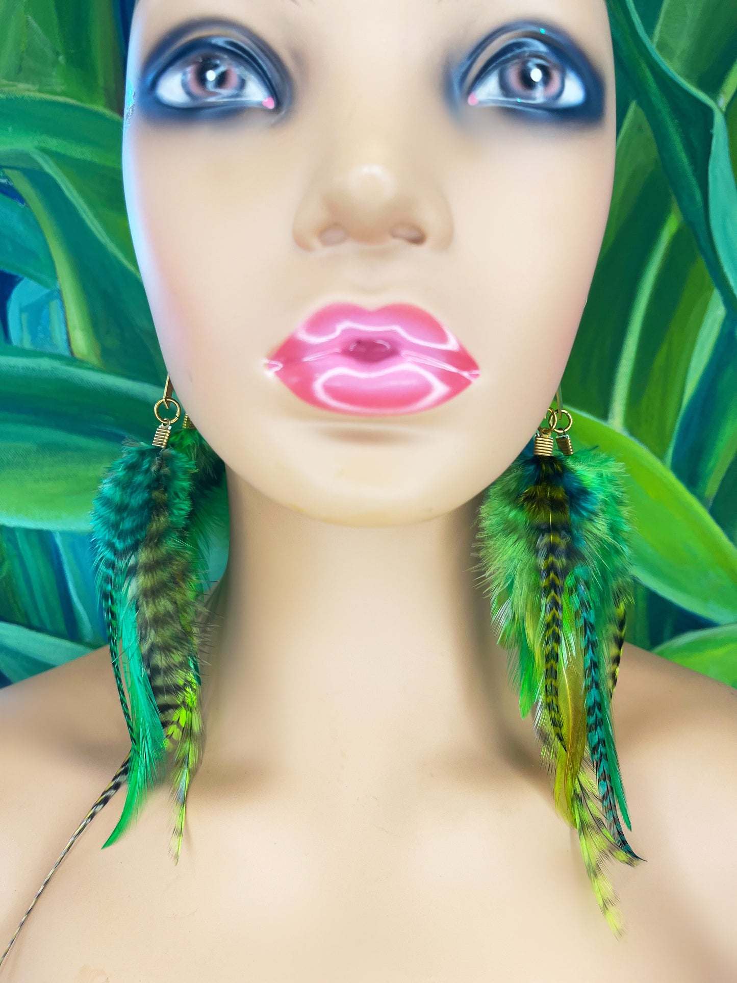 A mannequin head adorned with striking bohemian feather earrings that cascade in lush shades of green with black accent striping. The vibrant feathers stand out against the mannequin's neutral tone and the tropical leaf backdrop, embodying a wild and free-spirited aesthetic.