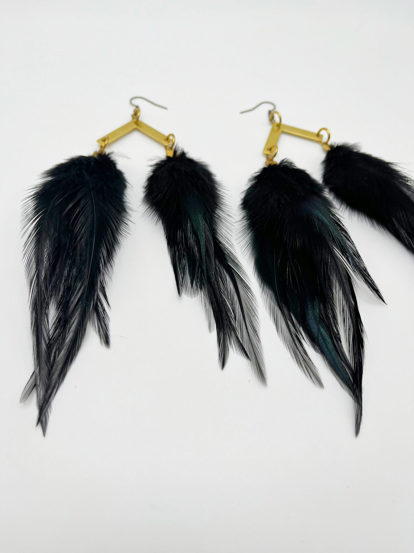 A pair of black feather earrings displayed against a plain white background. Each earring consists of several long, slender feathers descending from a gold-toned, diamond-shaped hook with a textured finish.