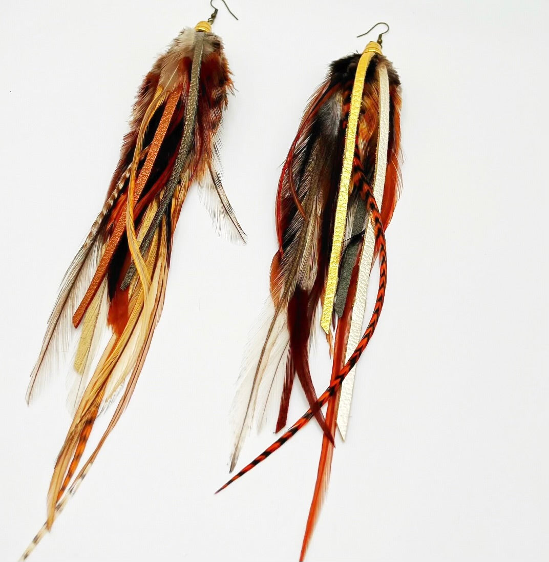 A pair of bohemian-style long earrings featuring a mix of natural brown feathers and hand-cut strips of metallic gold, bronze, and pewter leather. They are suspended from a gold-colored bronze bead cap and hypoallergenic ear wire, set against a white background, showcasing the earrings' layered and dynamic design.