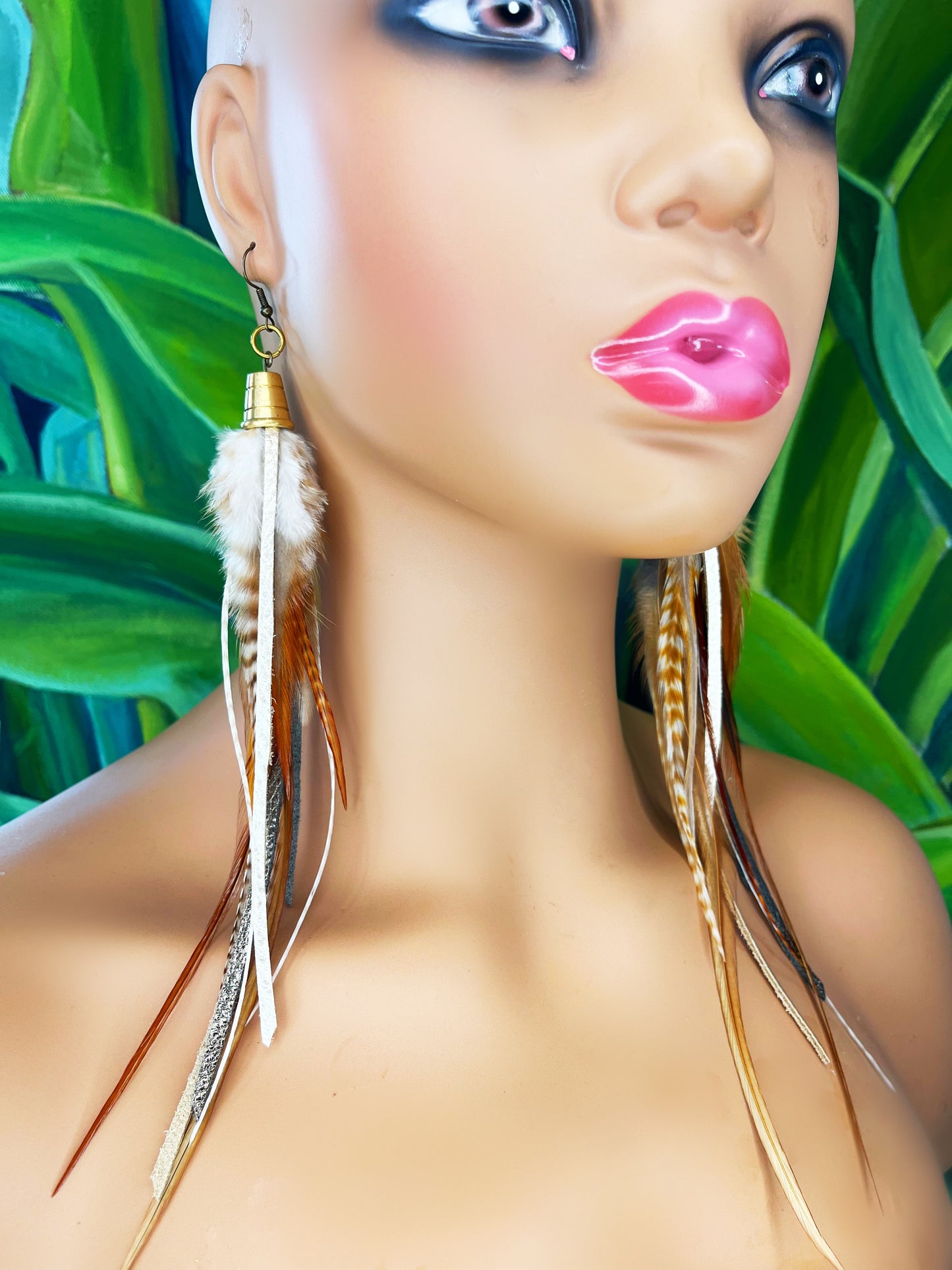 A model wearing long feather earrings in light brown and beige, with metallic bronze and pewter colored strips of leather, hanging from a gold colored cap pendant.