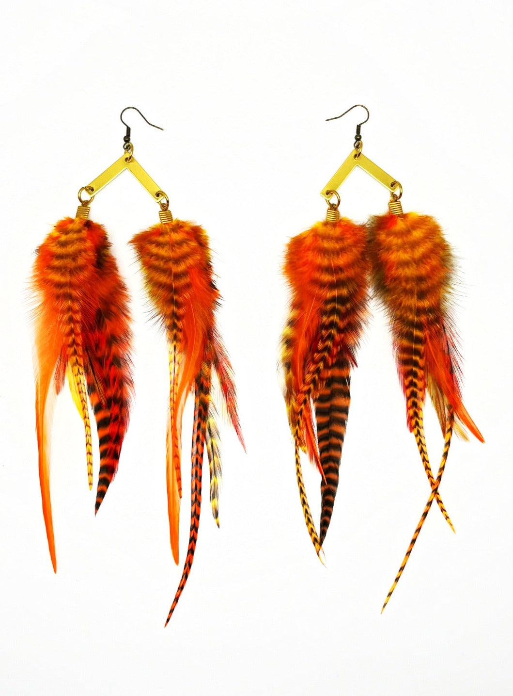 A pair of vibrant orange and striped feather earrings showcased against a white backdrop. The earrings feature a collection of feathers in fiery orange, with some having black and yellow striped patterns, giving them a wild and exotic appearance. They are attached to gold-toned, chevron-shaped hooks with a coil detail, which adds an elegant touch to their bold design. The arrangement of the feathers provides a sense of movement and a lush, full look.