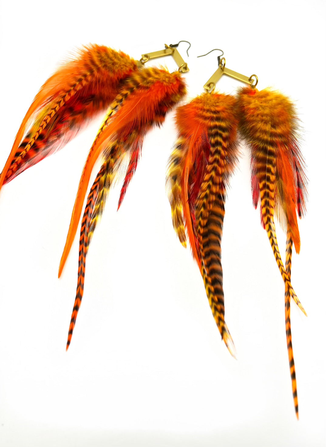 A pair of vibrant orange and striped feather earrings showcased against a white backdrop. The earrings feature a collection of feathers in fiery orange, with some having black and yellow striped patterns, giving them a wild and exotic appearance. They are attached to gold-toned, chevron-shaped hooks with a coil detail, which adds an elegant touch to their bold design. The arrangement of the feathers provides a sense of movement and a lush, full look.