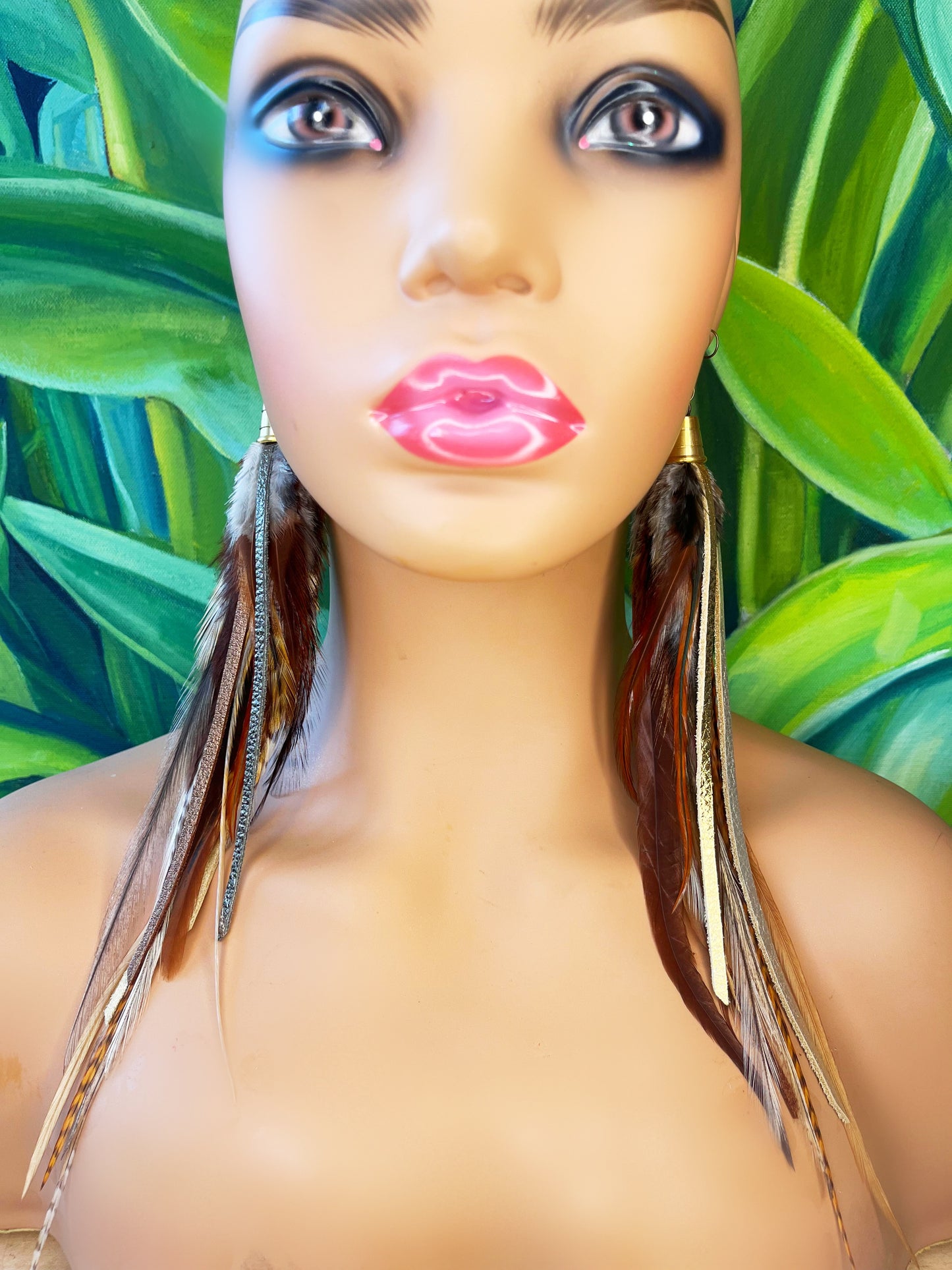 A mannequin head displays a pair of long bohemian earrings, featuring a combination of natural brown feathers and shimmering metallic leather strips in gold, bronze, and pewter. The earrings dangle from gold-colored bronze bead caps connected to hypoallergenic ear wires, set against a lush background of tropical green leaves, which accentuates the natural yet luxurious aesthetic of the jewelry.
