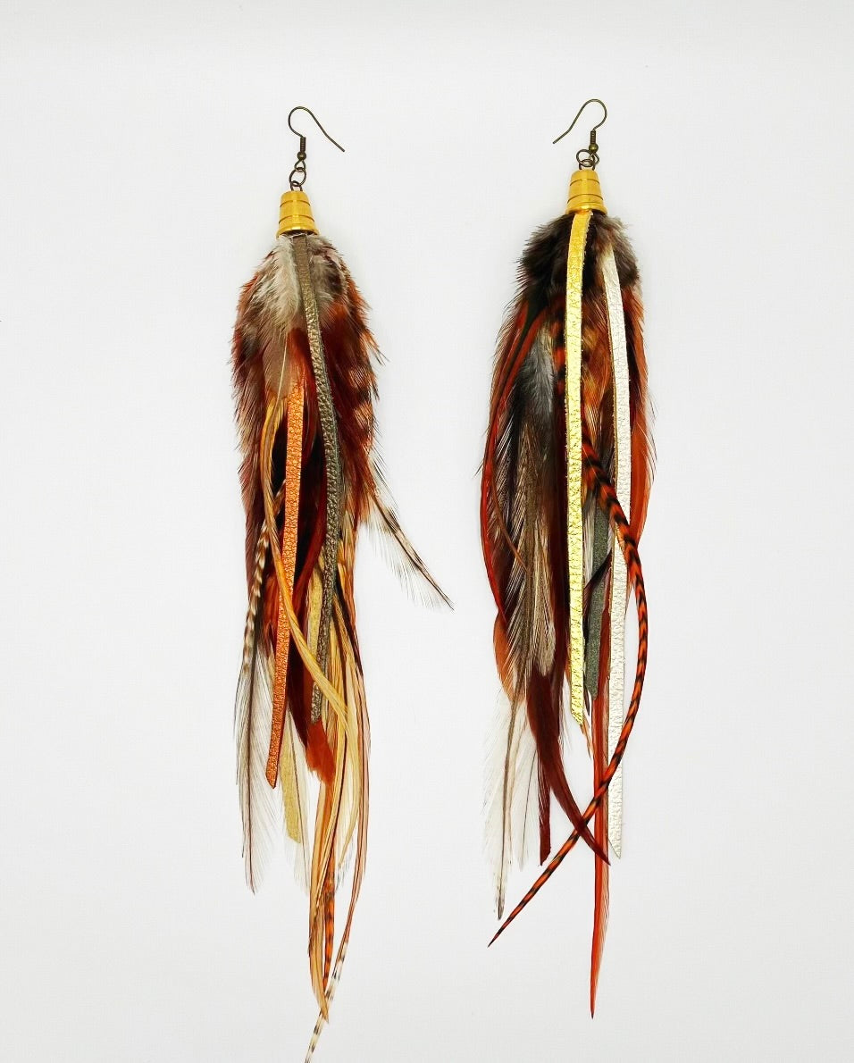 A pair of bohemian-style long earrings featuring a mix of natural brown feathers and hand-cut strips of metallic gold, bronze, and pewter leather. They are suspended from a gold-colored bronze bead cap and hypoallergenic ear wire, set against a white background, showcasing the earrings' layered and dynamic design.