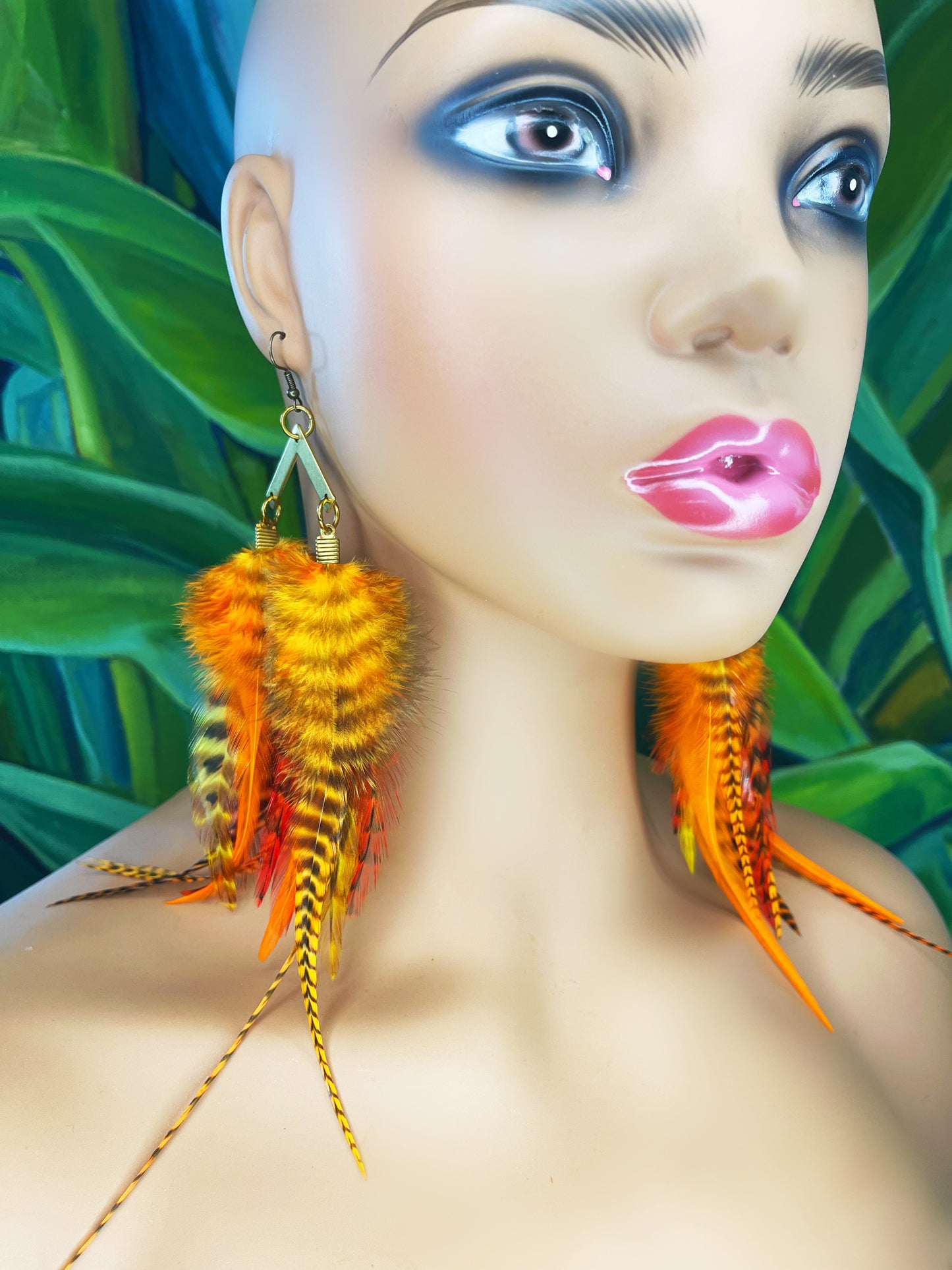 A mannequin wearing medium length bohemian feather earrings in bright golden orange shades, all feathers hang from a gold arrow pendant.