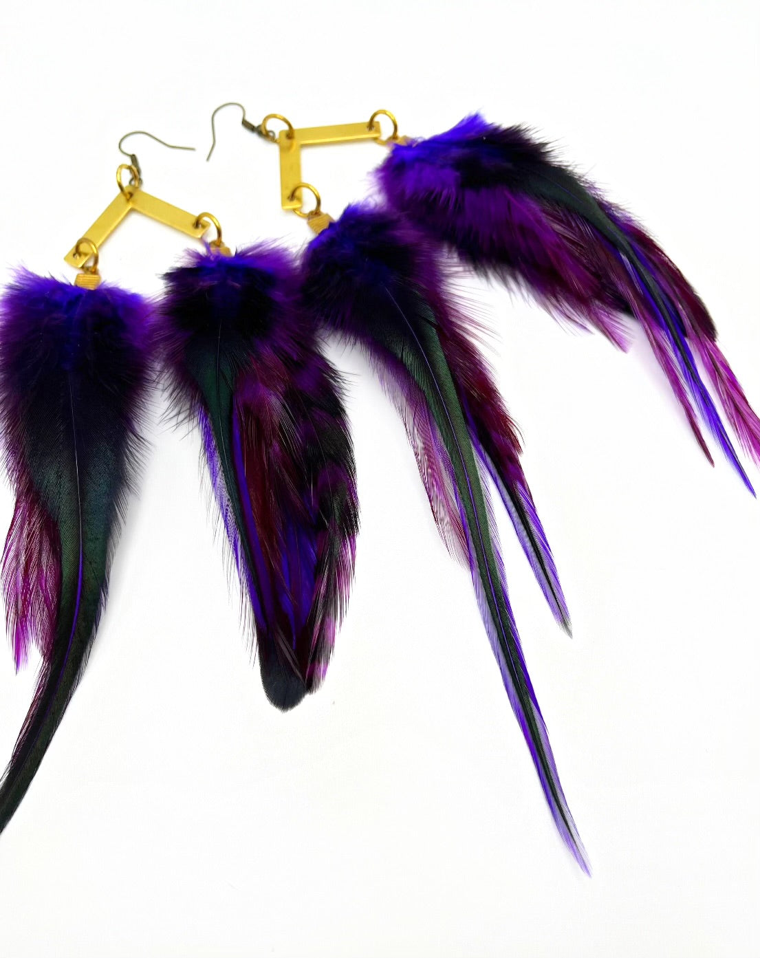 A pair of luxurious bohemian-style feather earrings, showcasing a rich gradient of purple to deep black. They are elegantly attached to gold geometric-shaped hardware, providing a sophisticated contrast against the white background. The feathers appear soft and fluffy, giving the earrings a dynamic and opulent look.