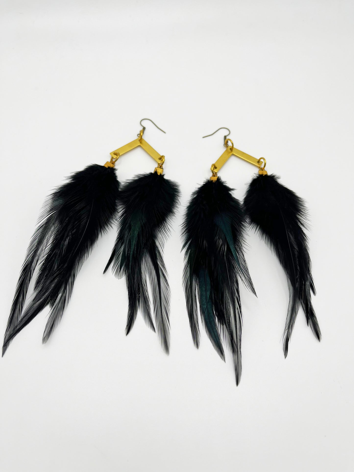 A pair of black feather earrings displayed against a plain white background. Each earring consists of several long, slender feathers descending from a gold-toned, diamond-shaped hook with a textured finish.