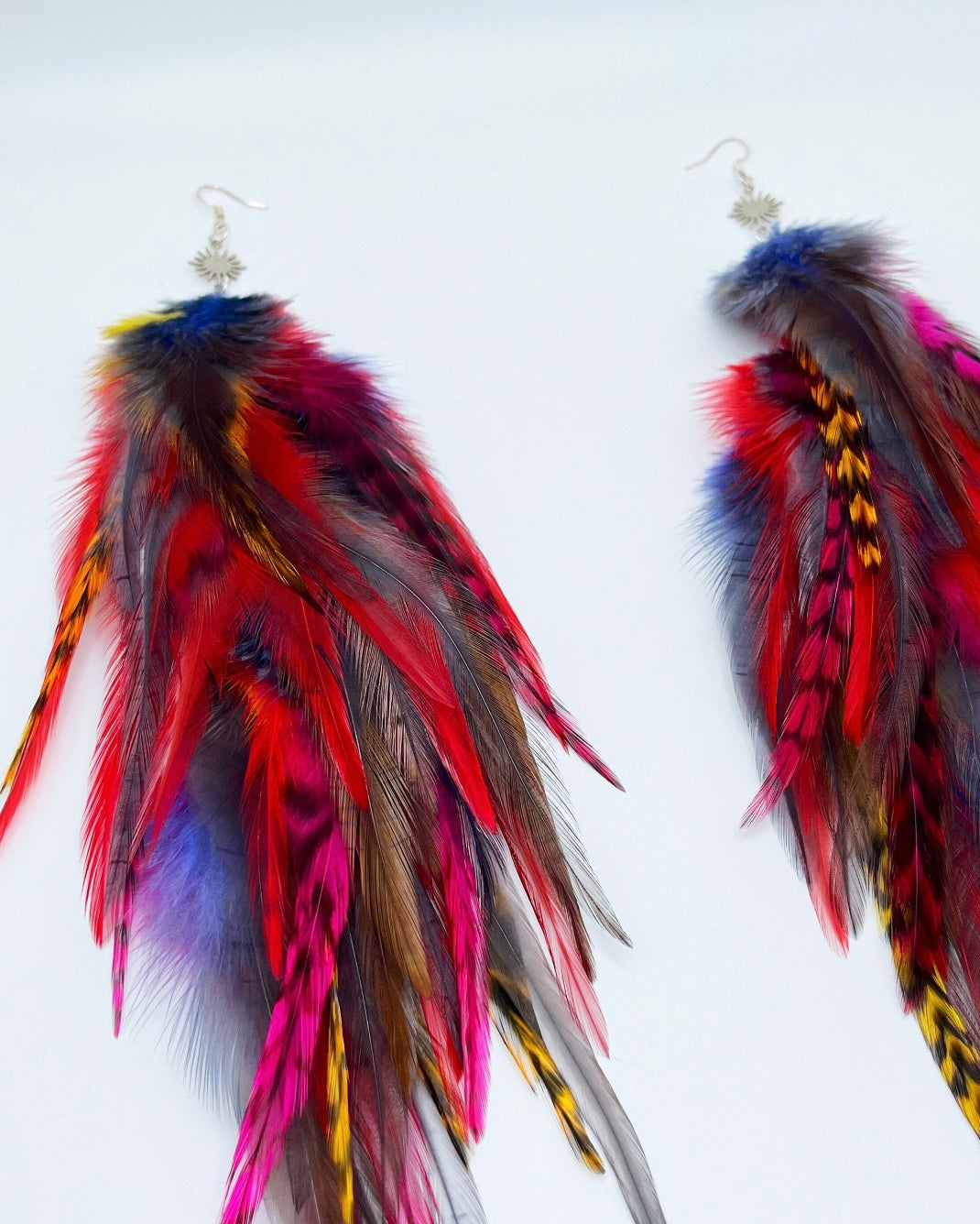 A pair of vibrant feather earrings displayed against a white background. The earrings are adorned with a variety of feathers in bold colors, including red, yellow, blue, and purple, with some natural brown feathers as well. Each earring has a silver fish hook style fastener with a small sunburst patterned charm attached to the top. The feathers are arranged in a cascading, slightly curved fashion, giving the earrings a dynamic and flowing appearance.