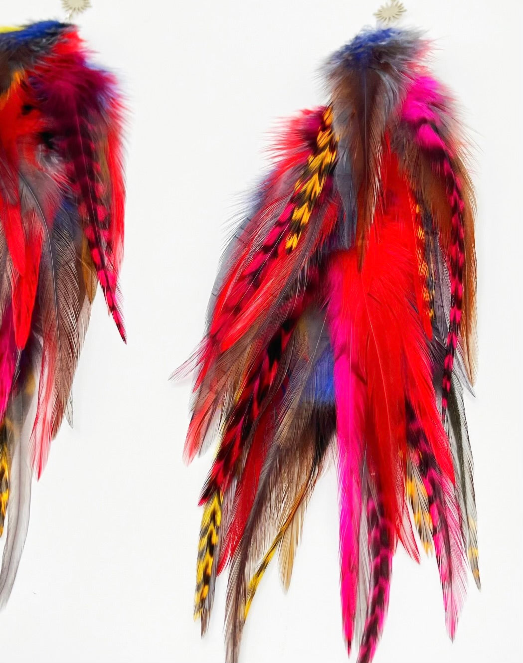 A pair of vibrant feather earrings displayed against a white background. The earrings are adorned with a variety of feathers in bold colors, including red, yellow, blue, and purple, with some natural brown feathers as well. Each earring has a silver fish hook style fastener with a small sunburst patterned charm attached to the top. The feathers are arranged in a cascading, slightly curved fashion, giving the earrings a dynamic and flowing appearance.