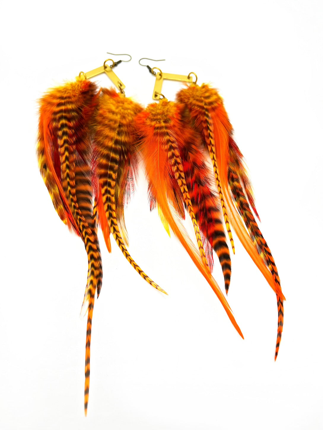 A pair of vibrant orange and striped feather earrings showcased against a white backdrop. The earrings feature a collection of feathers in fiery orange, with some having black and yellow striped patterns, giving them a wild and exotic appearance. They are attached to gold-toned, chevron-shaped hooks with a coil detail, which adds an elegant touch to their bold design. The arrangement of the feathers provides a sense of movement and a lush, full look.