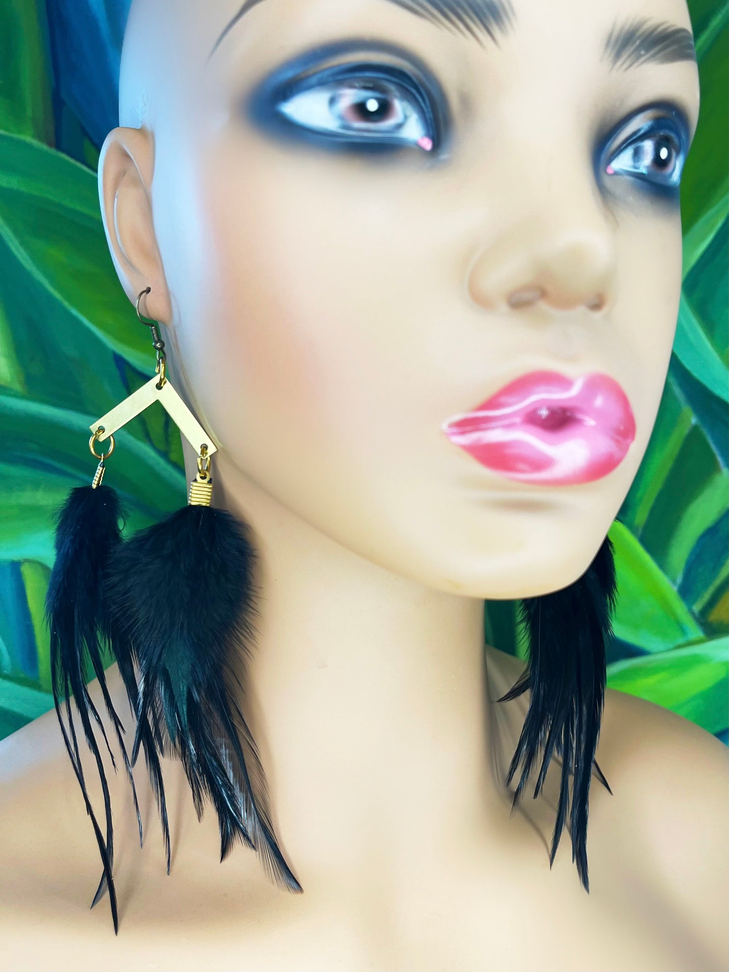 A close-up of a mannequin head wearing long black feather earrings. The earrings feature a gold-toned, geometric-shaped hook with a subtle hammered texture, leading to a cascade of sleek black feathers.