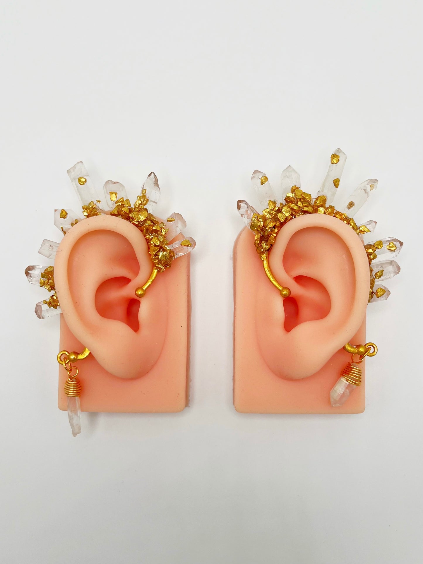 Clear quartz crystal ear cuffs with crushed gold accents, modelled on a pair of ears. A wire wrapped quartz stone hangs from the bottom of the cuffs behind the ears.