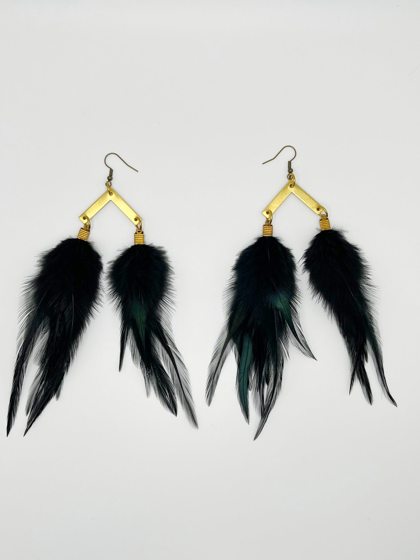 A pair of black feather earrings displayed against a plain white background. Each earring consists of several long, slender feathers descending from a gold-toned, diamond-shaped hook with a textured finish.