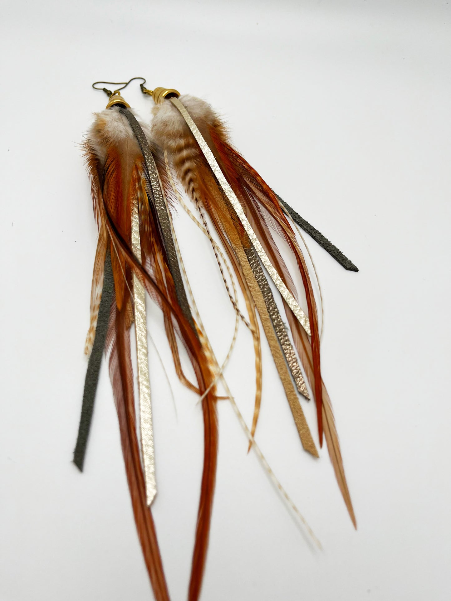 Long feather earrings in light brown and beige, with metallic bronze and pewter colored strips of leather, hanging from a gold colored cap pendant.