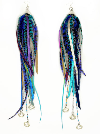 A pair of long, dangling feather and ribbon earrings with silver fish-shaped charms, featuring a mix of blue, teal, purple, and black feathers on a white background.