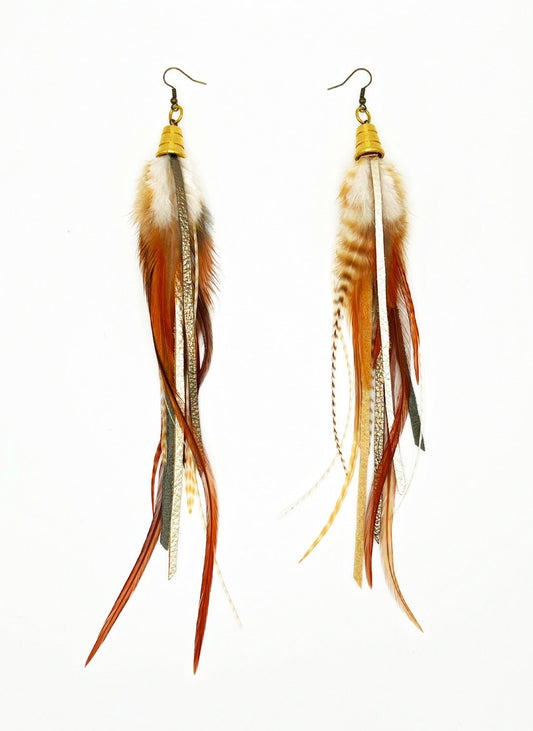 Long feather earrings in light brown and beige, with metallic bronze and pewter colored strips of leather, hanging from a gold colored cap pendant.