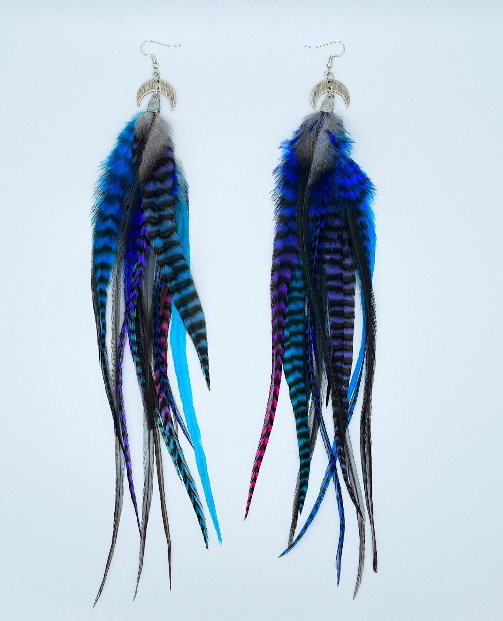 A pair of long bohemian feather earrings, featuring a striking array of hand-dyed grizzly feathers in vibrant shades of blue, purple, and teal, complemented by natural dark feathers. The feathers are attached to a silver crescent moon pendant connected to silver ear wires, creating a layered and flowing design against a white background.