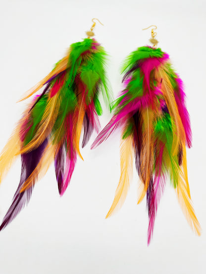 Long bohemian feather earrings in bright green, pink, purple and orange tones, hanging from a gold sun pendant. 