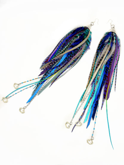 A pair of long, dangling feather and ribbon earrings on a plain white background. The earrings feature a mix of blue, purple, teal, and silver feathers with silver wire loops, silver fishhook ear wires, and small silver heart charms at the ends.
