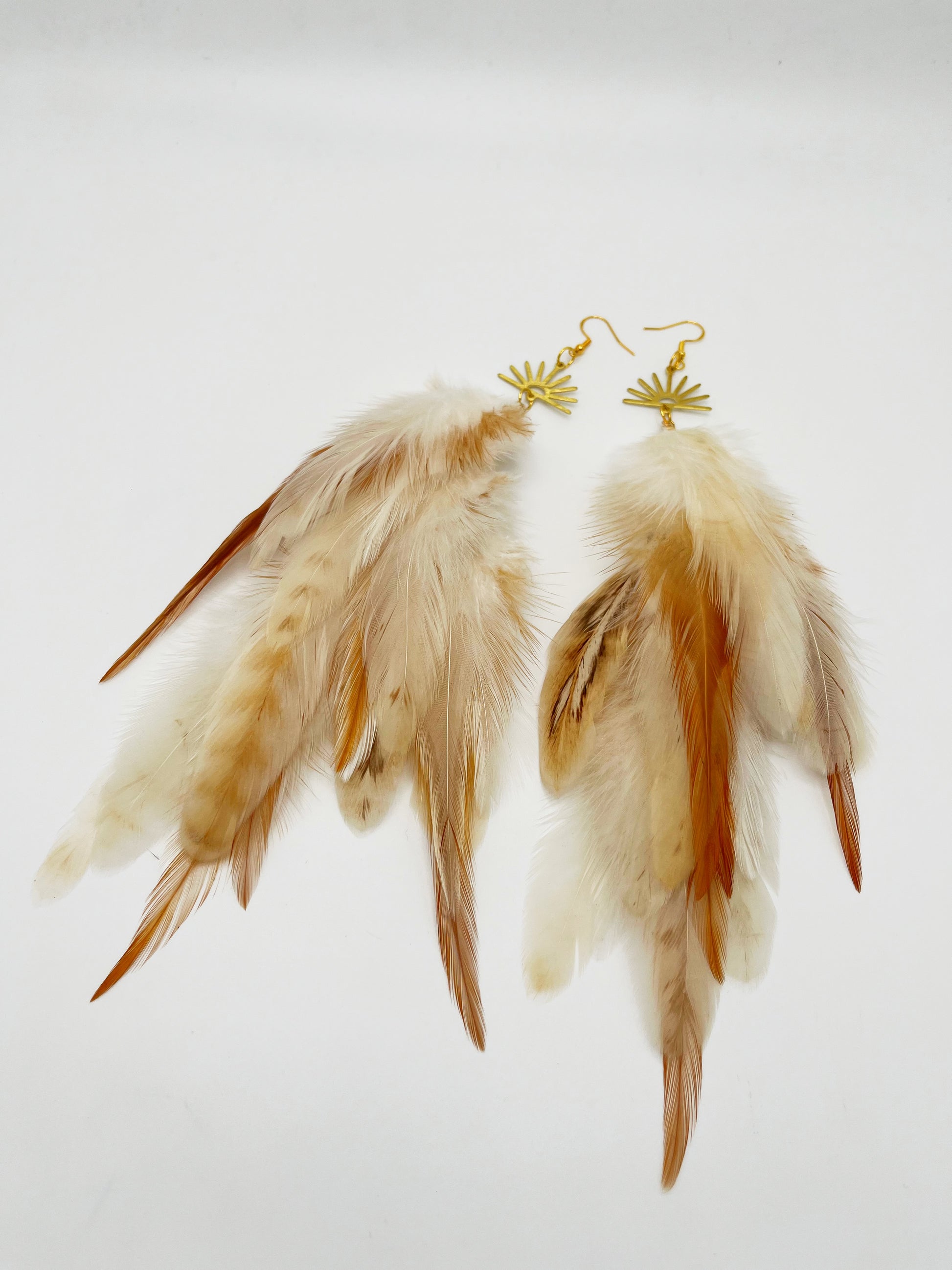 Long bohemian feather earrings in cream and beige tones, hanging from a gold sun pendant. 