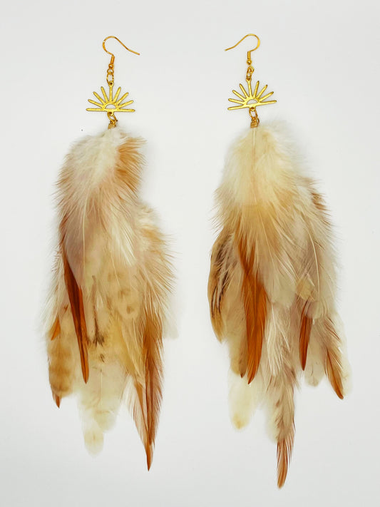 Long bohemian feather earrings in cream and beige tones, hanging from a gold sun pendant. 