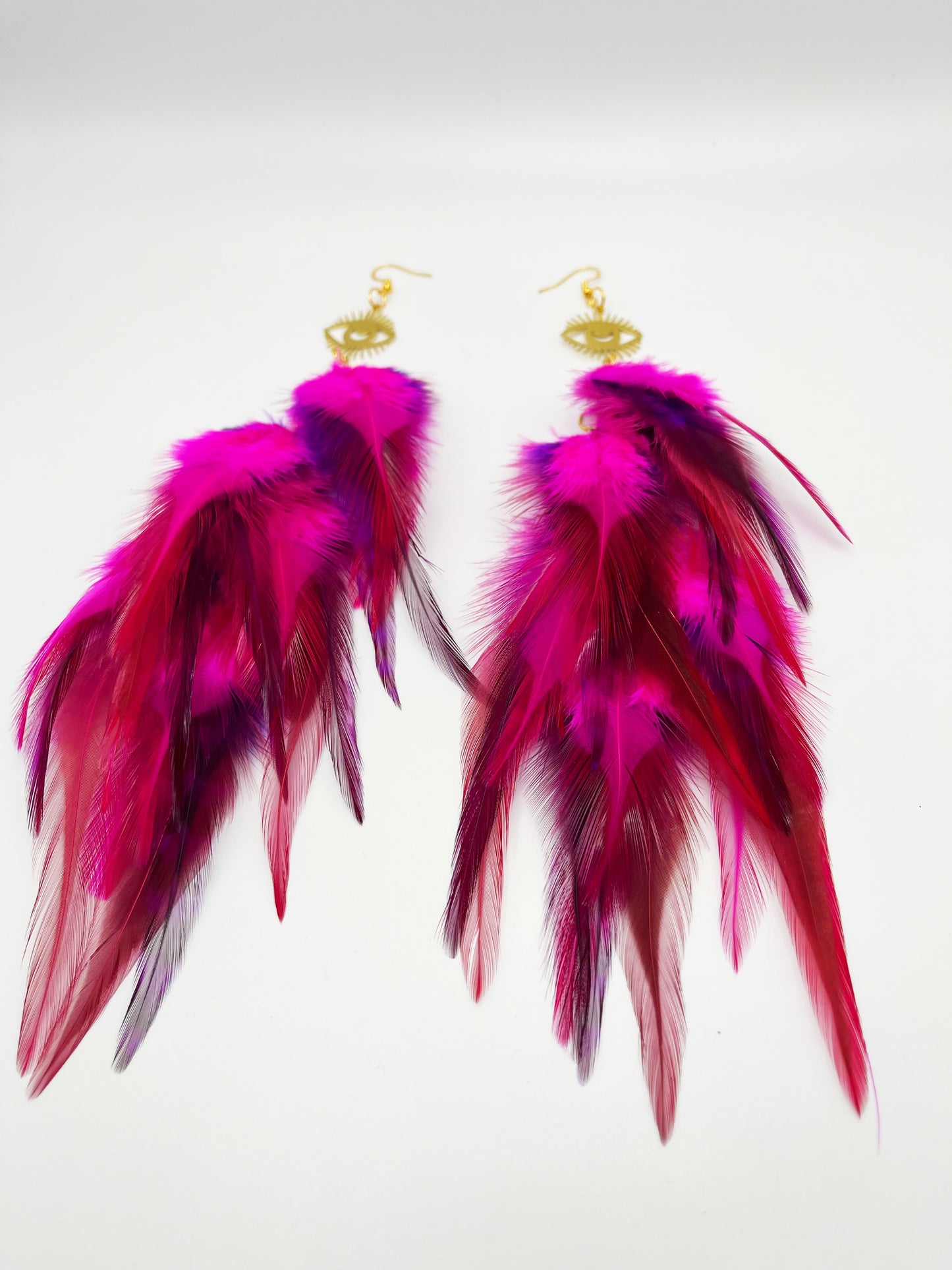 Long bohemian feather earrings in bright pink colors, hanging from a gold eye pendant. 