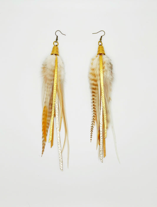 White, cream and beige feather earrings with strips of metallic gold and pewter leather, hanging from a gold colored bead pendant. 