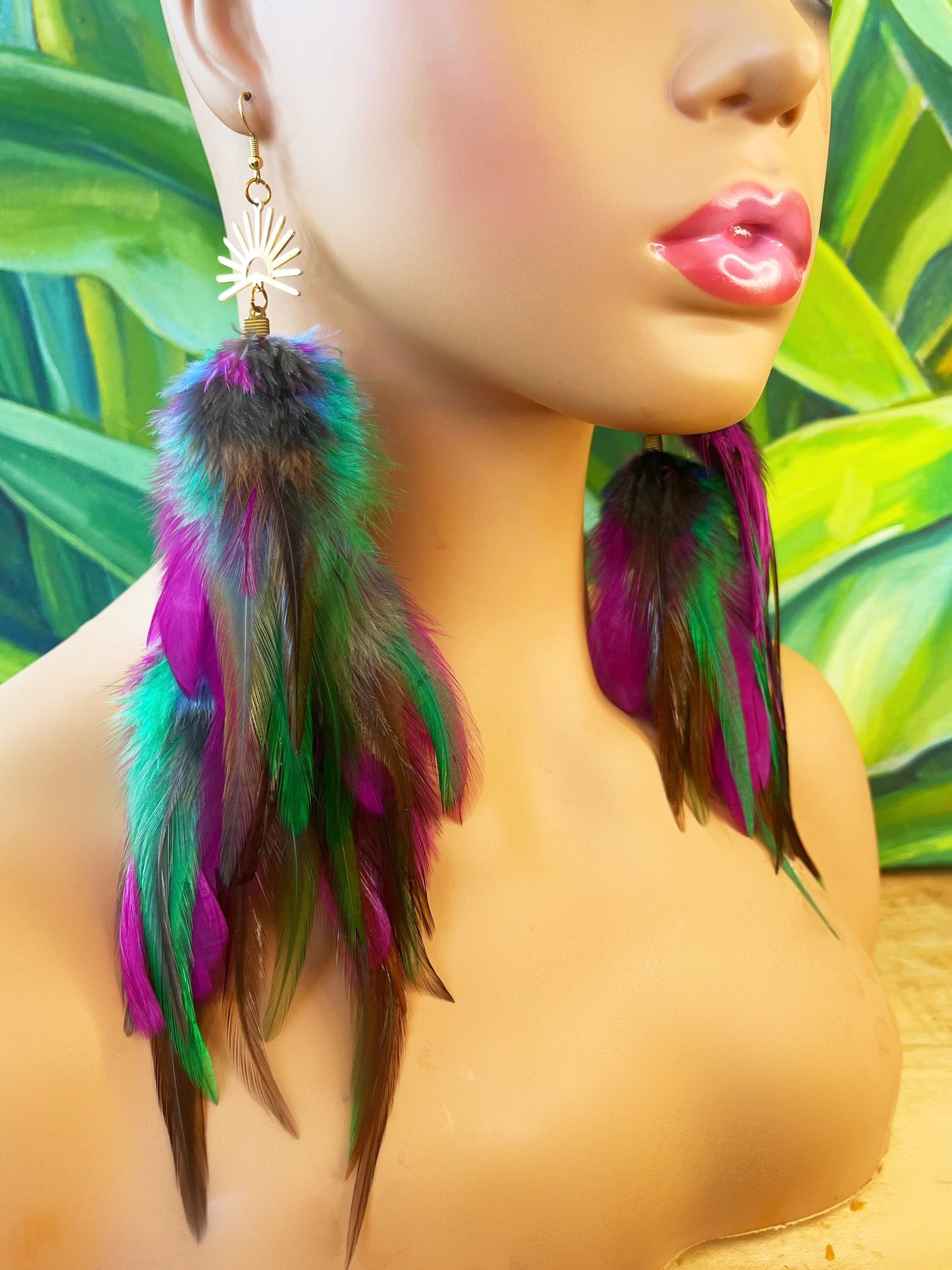 Vibrant Mardi Gras-inspired long feather earrings in purple, green, and pink, with a gold sunburst pendant, displayed on a mannequin against a tropical leaf background.