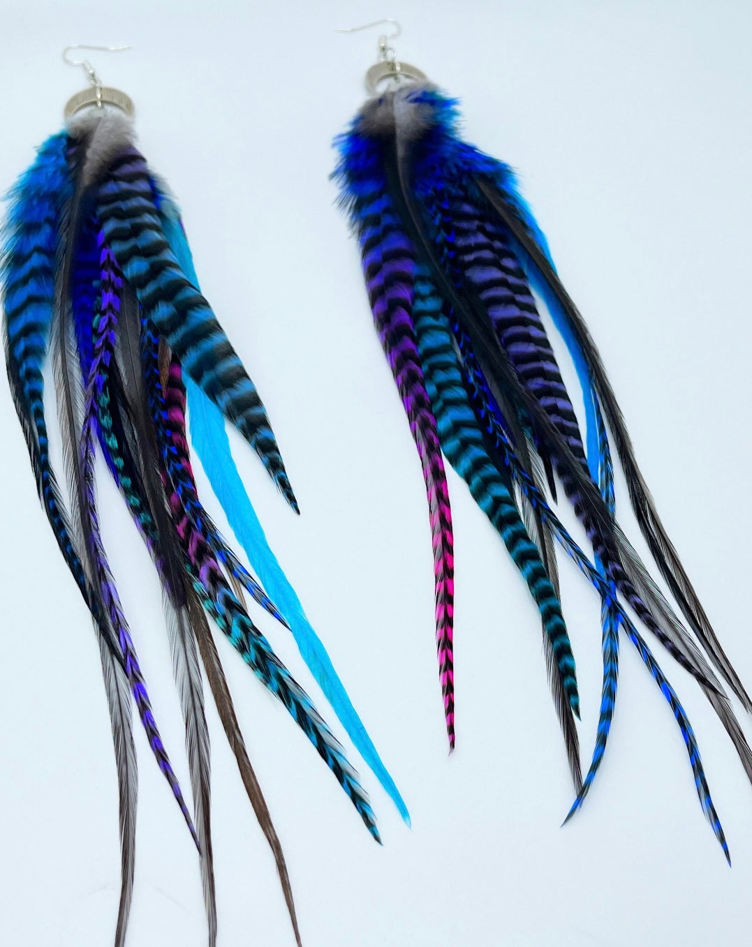 A pair of long bohemian feather earrings, featuring a striking array of hand-dyed grizzly feathers in vibrant shades of blue, purple, and teal, complemented by natural dark feathers. The feathers are attached to a silver crescent moon pendant connected to silver ear wires, creating a layered and flowing design against a white background.