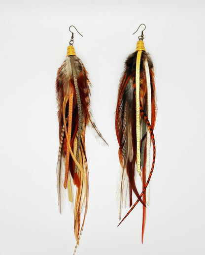  A pair of bohemian-style long earrings featuring a mix of natural brown feathers and hand-cut strips of metallic gold, bronze, and pewter leather. They are suspended from a gold-colored bronze bead cap and hypoallergenic ear wire, set against a white background, showcasing the earrings' layered and dynamic design.