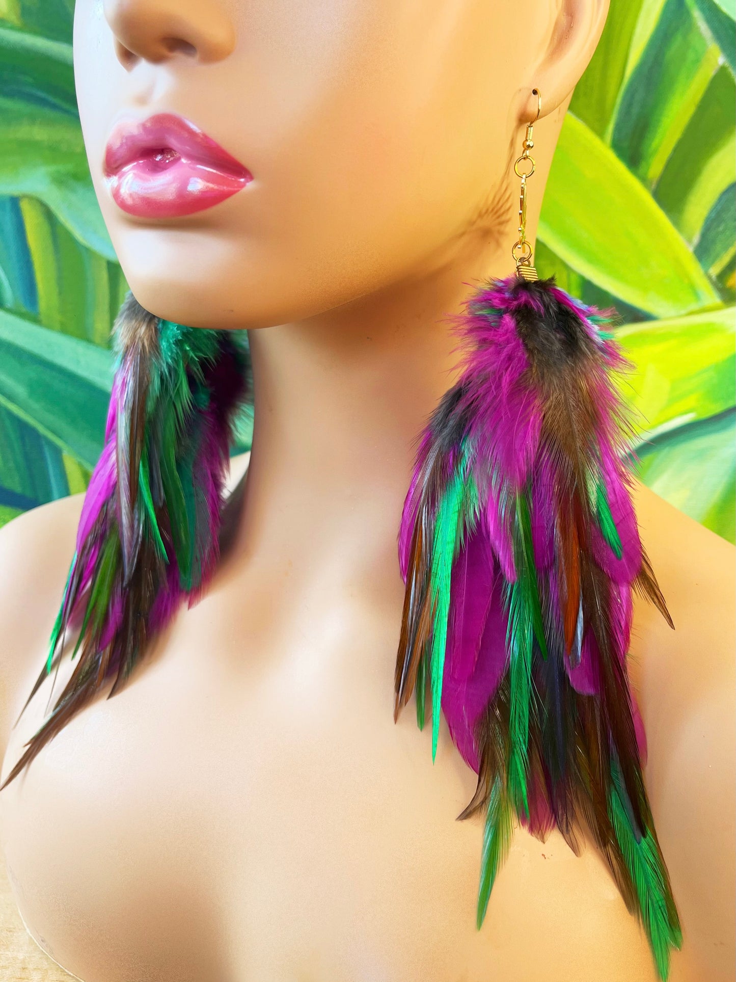 Vibrant Mardi Gras-inspired long feather earrings in purple, green, and pink, with a gold sunburst pendant, displayed on a mannequin against a tropical leaf background.