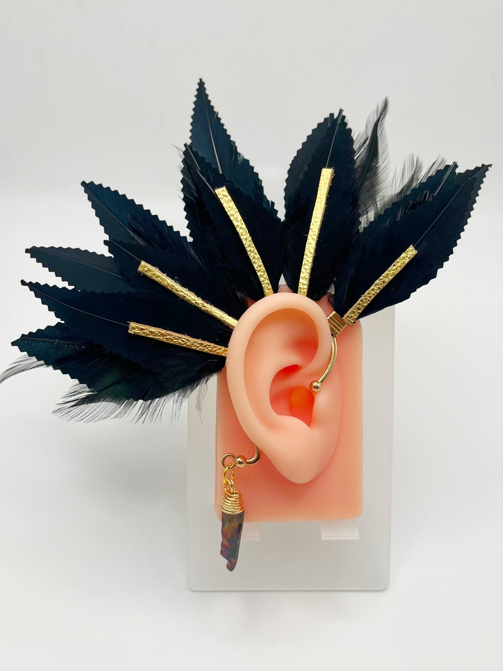 Structured black feather ear cuff with gold leather accents modeled on an ear. A wire wrapped quartz hangs from the bottom of the cuff behind the ear.