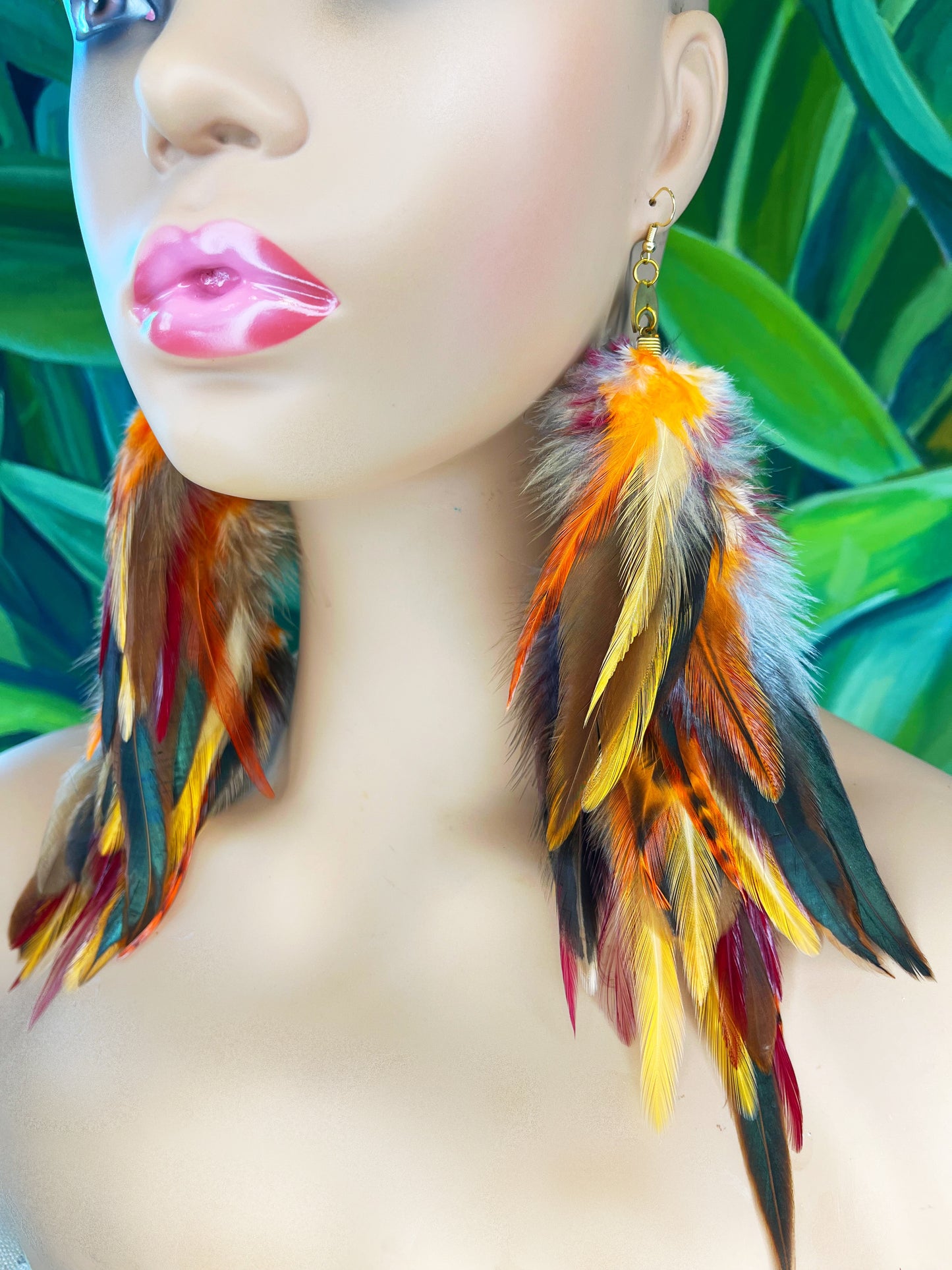 Mannequin showcasing 'Autumn's Gaze' Long Feather Earrings with a blend of orange, red, and yellow feathers and a gold link chain against a tropical leaf background.