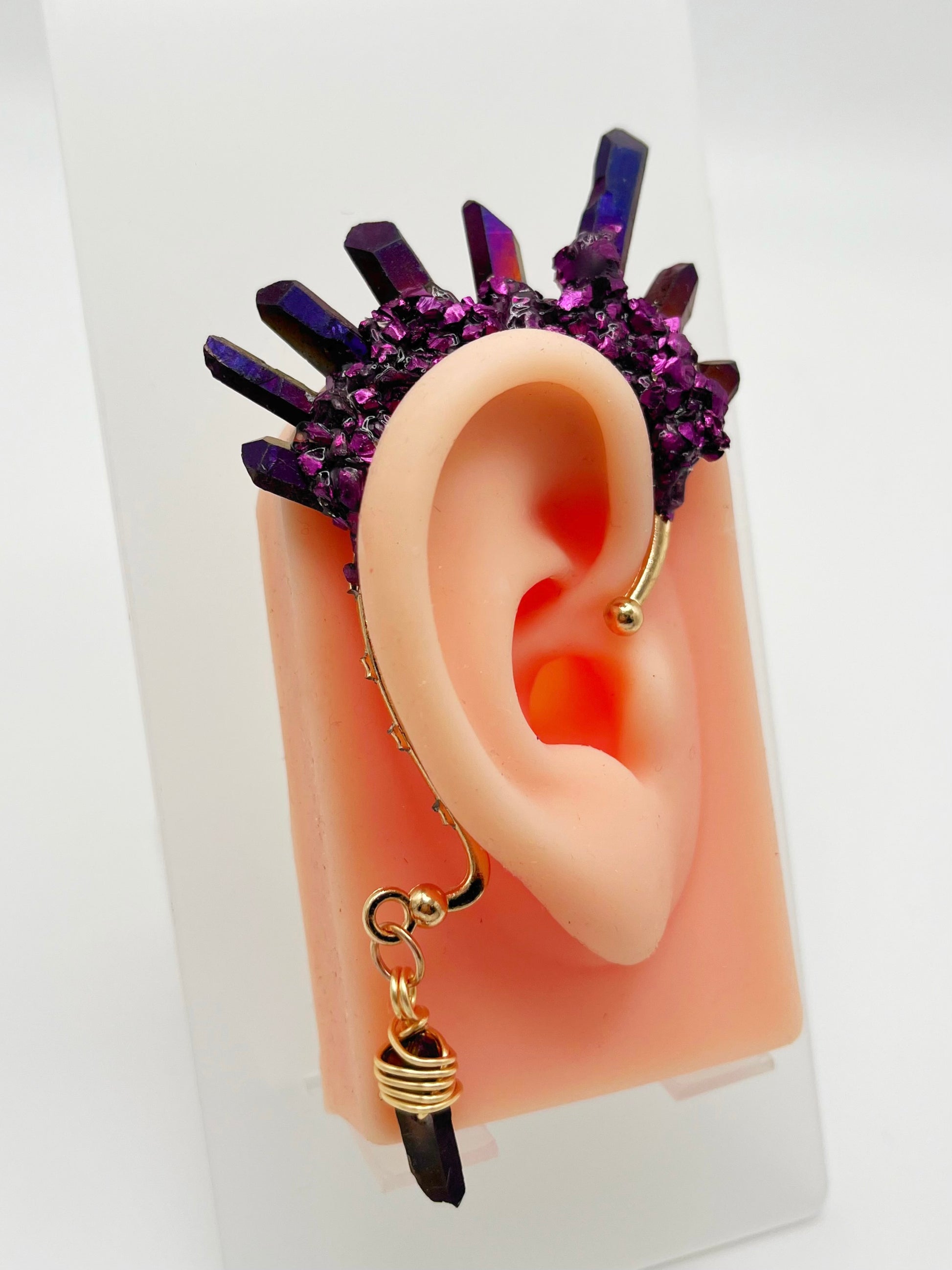Purple titanium coated quartz ear cuff with metallic purple accents, modeled on an ear. A wire wrapped quartz stone is attached to the bottom of the cuff that hangs behind the ear.