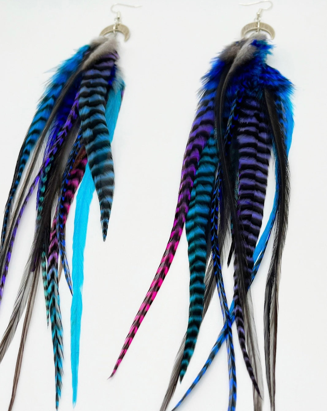 A pair of long bohemian feather earrings, featuring a striking array of hand-dyed grizzly feathers in vibrant shades of blue, purple, and teal, complemented by natural dark feathers. The feathers are attached to a silver crescent moon pendant connected to silver ear wires, creating a layered and flowing design against a white background.