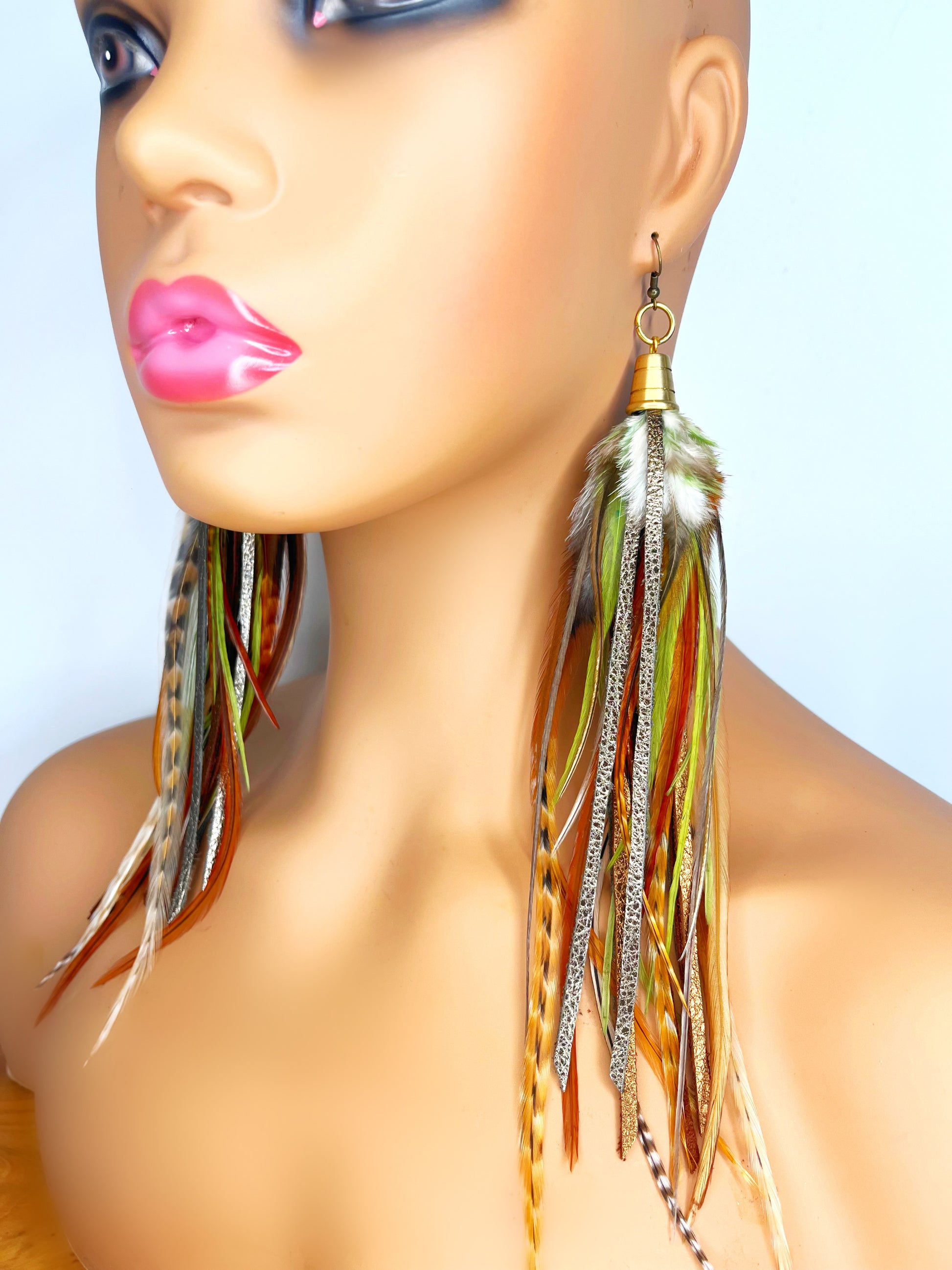 Mannequin wearing long feather earrings in green and brown tones, with hand cut metallic leather. All feathers and leather hang from a gold colored bead cap. 