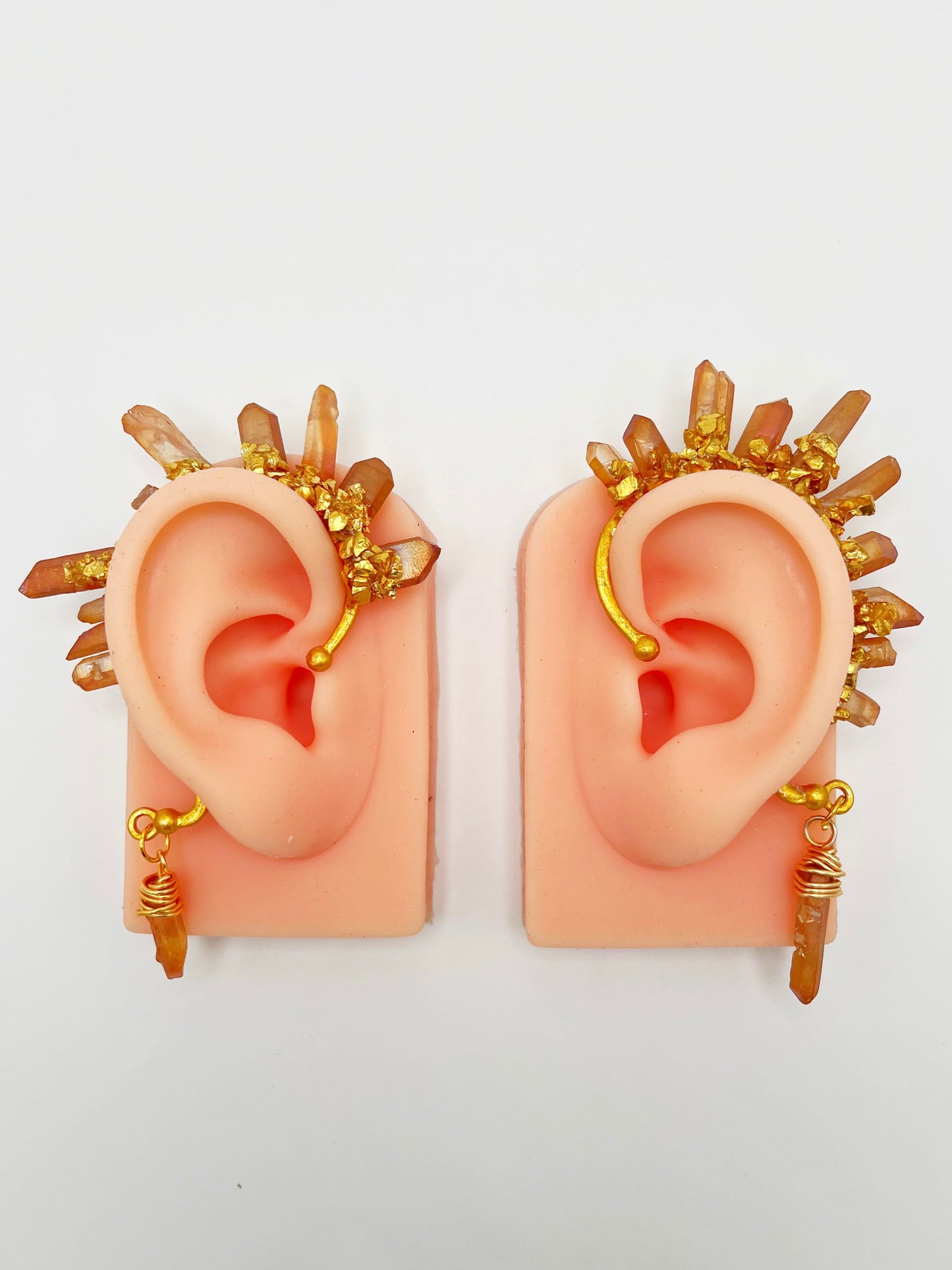 A pair of orange citrine crystal ear cuffs with crushed gold accents, modeled on a pair of ears. A wire wrapped stone hangs from the bottom of each cuff.