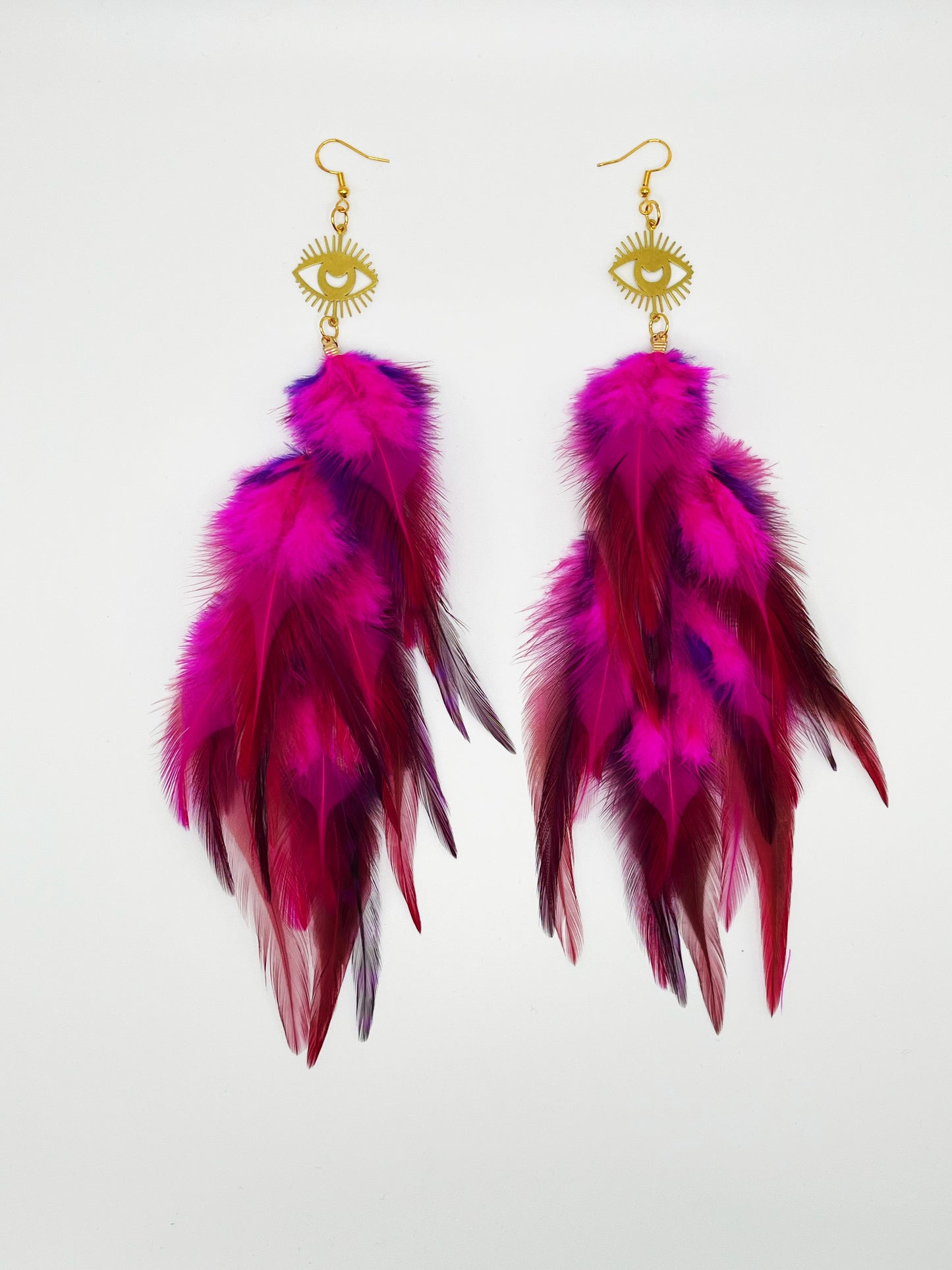 Long bohemian feather earrings in bright pink colors, hanging from a gold eye pendant. 