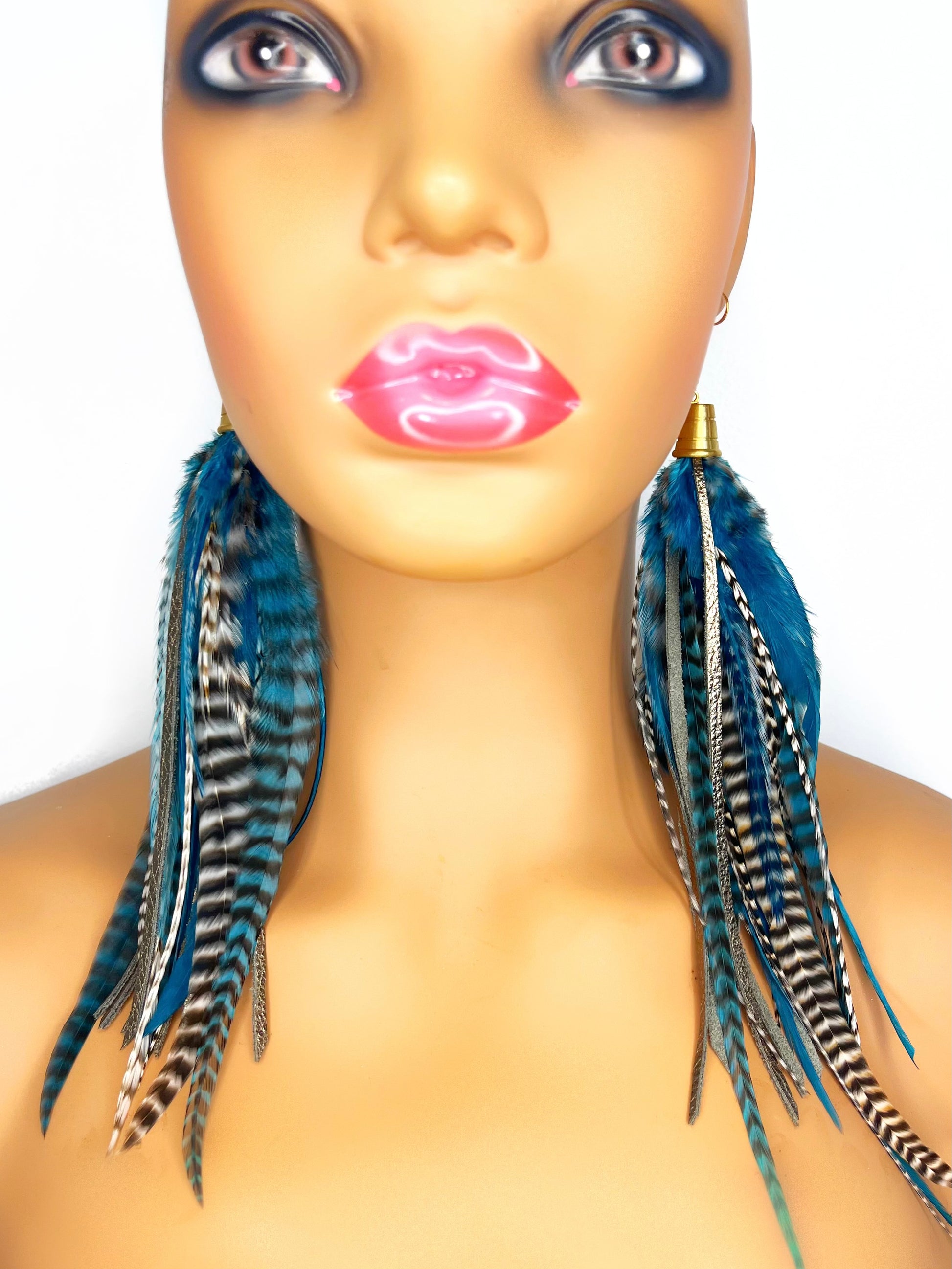 Mannequin wearing long teal rooster feather earrings with strips of metallic bronze leather. Feathers and leather hang from under a gold colored pendant.