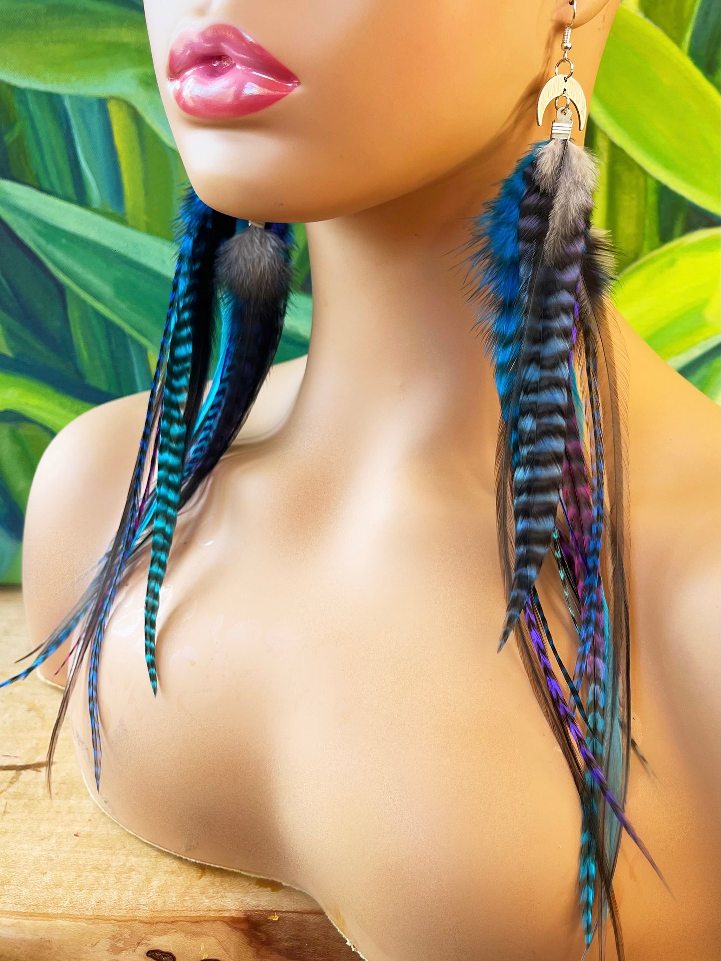 A mannequin head is adorned with long bohemian feather earrings featuring an ombre transition from deep blue to purple and pink grizzly feathers. The feathers cascade from a silver crescent moon pendant on silver ear wires, creating a layered look that complements the tropical greenery in the background