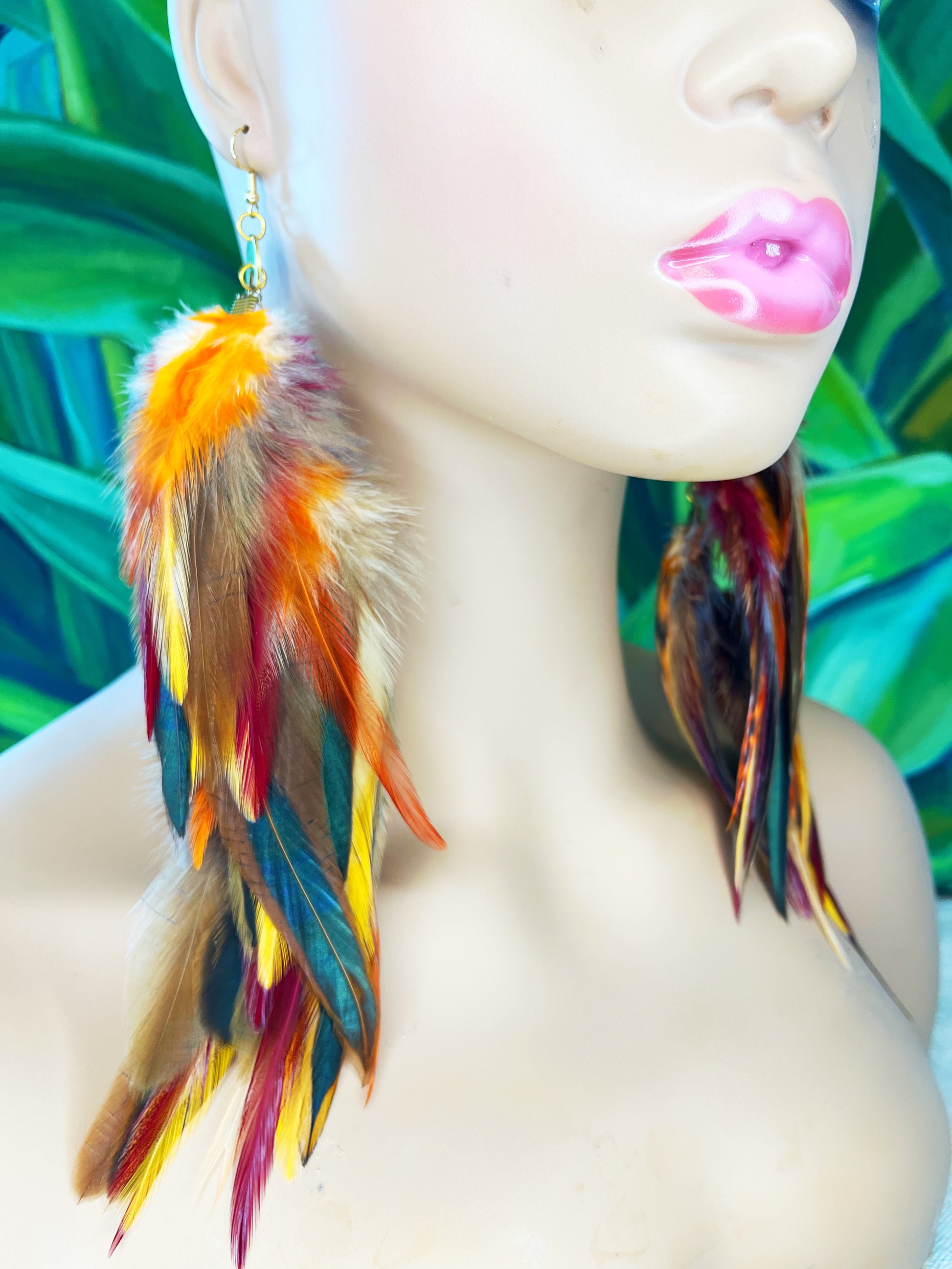 Mannequin showcasing 'Autumn's Gaze' Long Feather Earrings with a blend of orange, red, and yellow feathers and a gold link chain against a tropical leaf background.