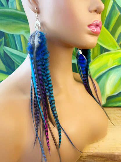 A mannequin head is adorned with long bohemian feather earrings featuring an ombre transition from deep blue to purple and pink grizzly feathers. The feathers cascade from a silver crescent moon pendant on silver ear wires, creating a layered look that complements the tropical greenery in the background